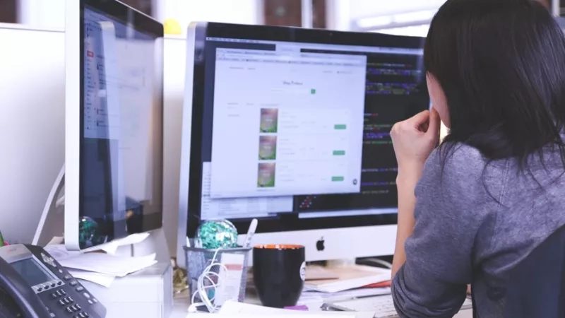 A woman sitting at a desk in front of two computers with a mug in between.