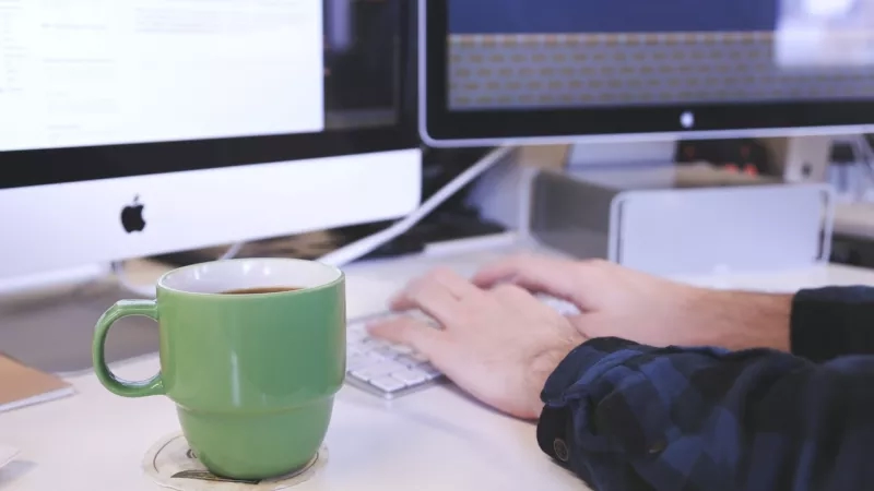 A person typing on a keyboard next to a green mug of coffee.