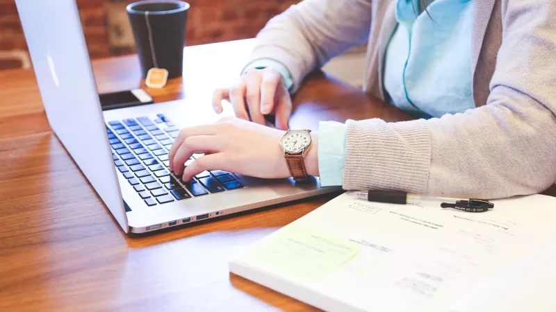 A woman is typing on a laptop while a notebook is on the desk in front of her.