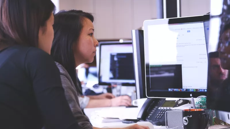 Two people are working on the computer in the office with many other people in the background.
