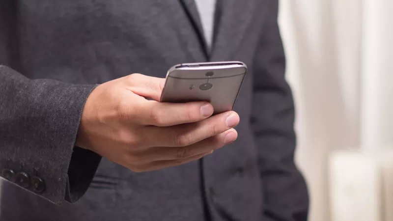 A man in a business suit is holding a mobile phone, possibly an iPhone or Android.
