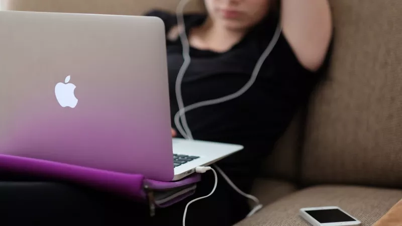 A woman with headphones is sitting on a couch with a laptop and a cell phone on her lap.