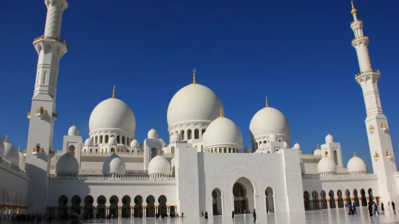 A large white building with multiple domes and a blue sky in the background, likely a mosque.