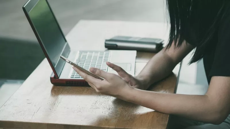 A woman is sitting on a table and using her phone while looking at her laptop.