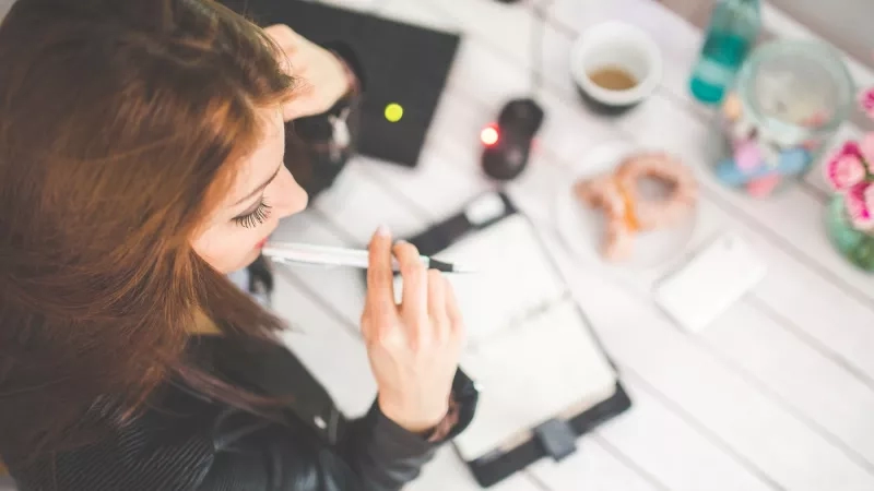 Woman in office, hand on desk with pen and notepad, and a cup of coffee nearby.