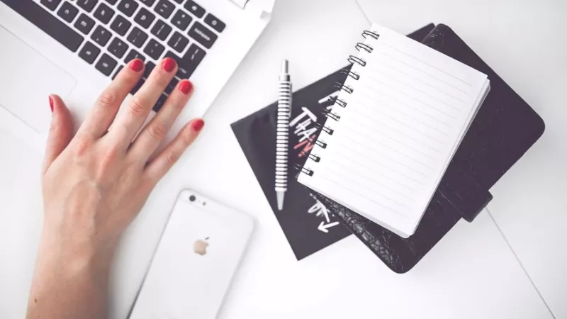Woman typing on laptop with smartphone, notepad and pen on desk