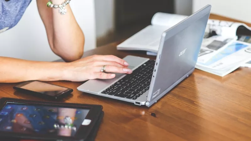 A woman uses a laptop computer at a desk, with a smartphone and tablet nearby.