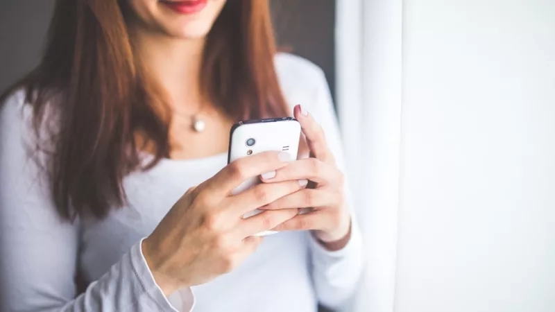 Young woman using her smartphone and smiling at the camera, standing against a white curtain.