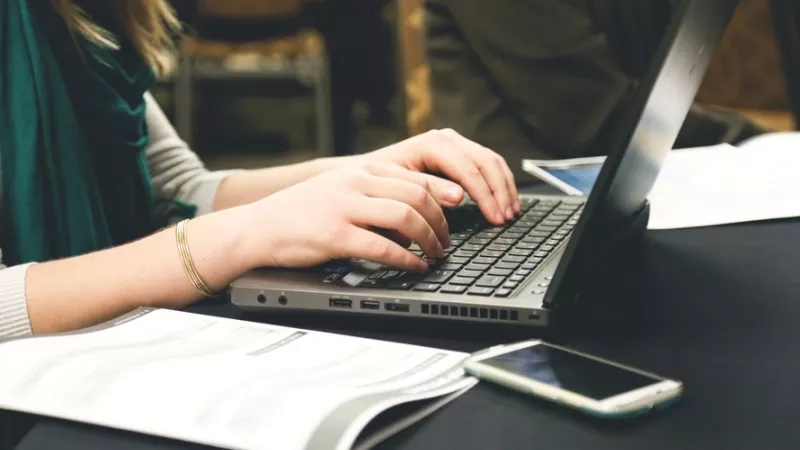 Woman typing on a laptop with a book and a cell phone on a table.