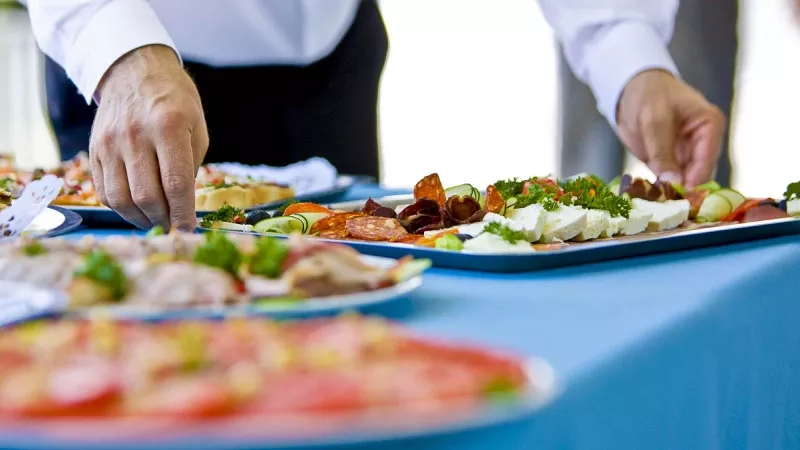 A person is placing food on a plate on a table covered with a blue cloth.