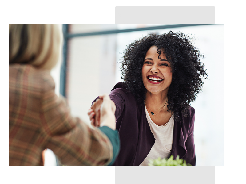 A female is shaking hands with a smiling female with curly hair in an office environment.