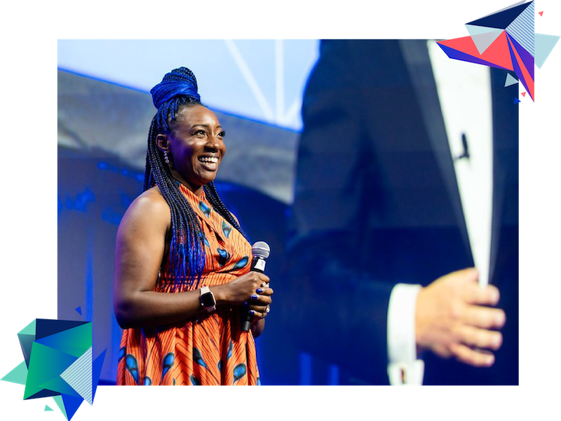 A woman with braided hair is standing on a stage holding a microphone and smiling at the camera.