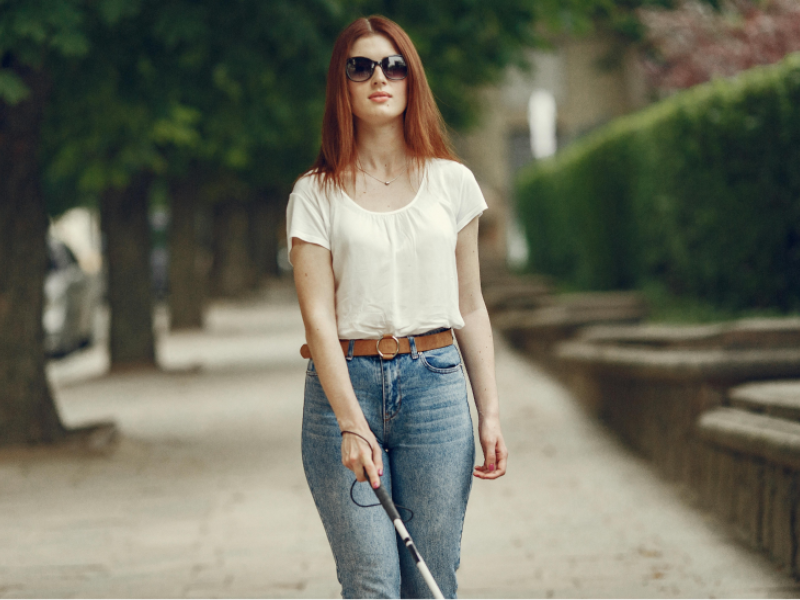 A woman in sunglasses and a white blouse holding a cane walks down the sidewalk.