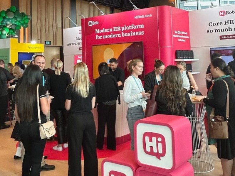 Several people are gathered around a HiBob stand at an event with a red carpet and balloons.
