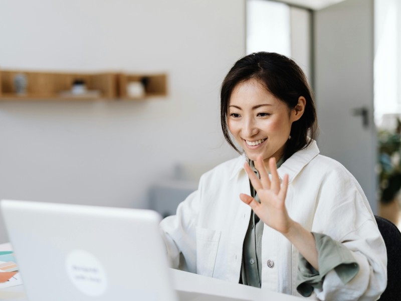 A woman in a white coat waves at her laptop in a modern office setting.