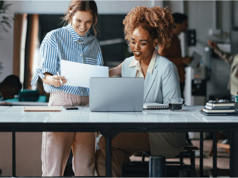 Two women are laughing and reviewing documents with a laptop in front of them in a casual office environment.