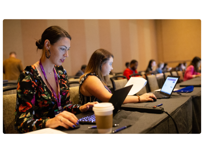 Two women in a conference room working on laptops with a cup of coffee on the table.