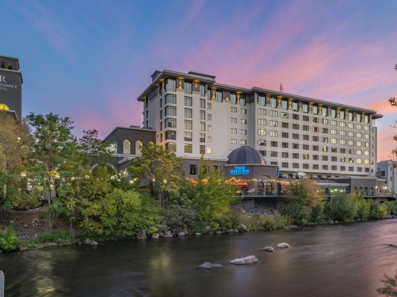 The Shore Hotel by the river at dusk, featuring trees, lights, and a vibrant sky.