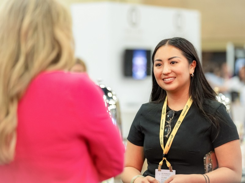 Woman in black shirt and yellow lanyard talking to woman in pink sweater at a trade show booth