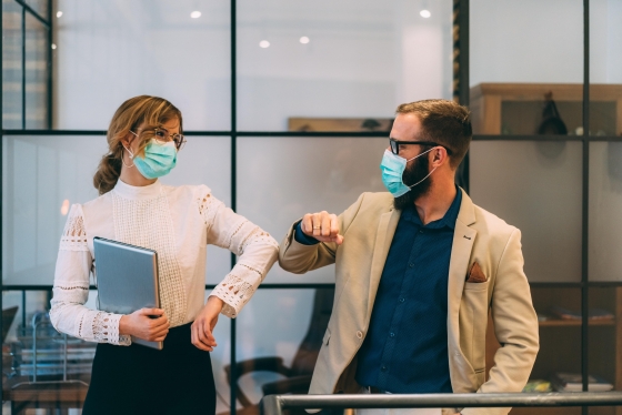 A man and woman wearing masks standing in an office while shaking hands and smiling.