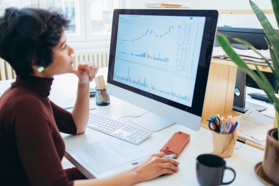 Woman looks at data on computer with pens, pencils, and coffee on desk next to her.