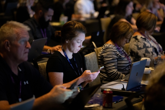 People at a conference, some using their phones and others using laptops, with coffee and a can nearby.
