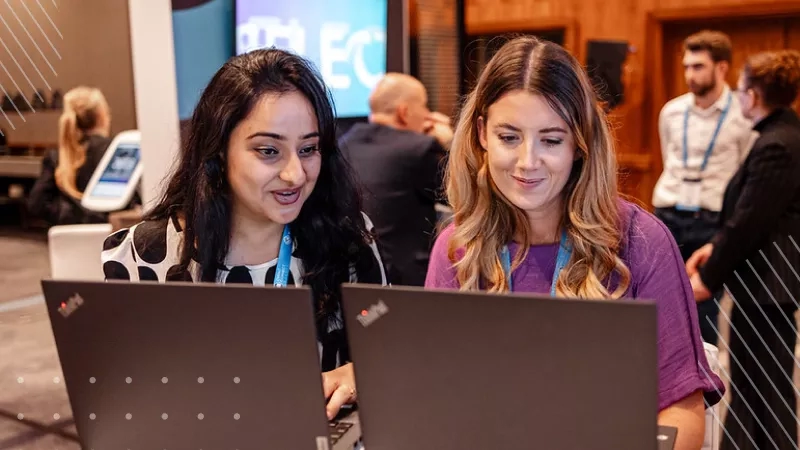 Two ladies using their laptops at an event with other people in the background.